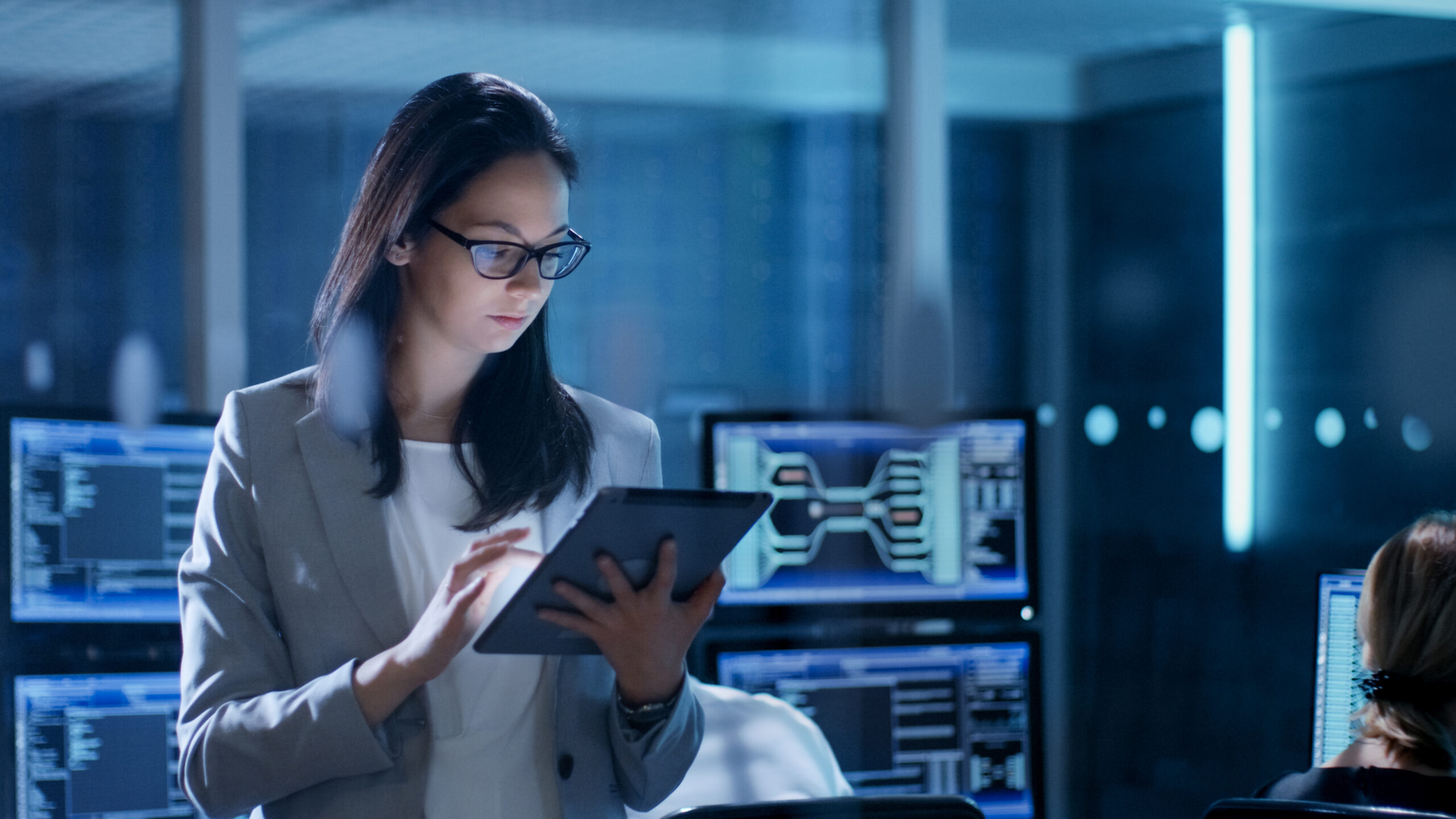 A woman in glasses uses a tablet in a tech workspace, surrounded by computer monitors displaying code, highlighting a focused work environment.