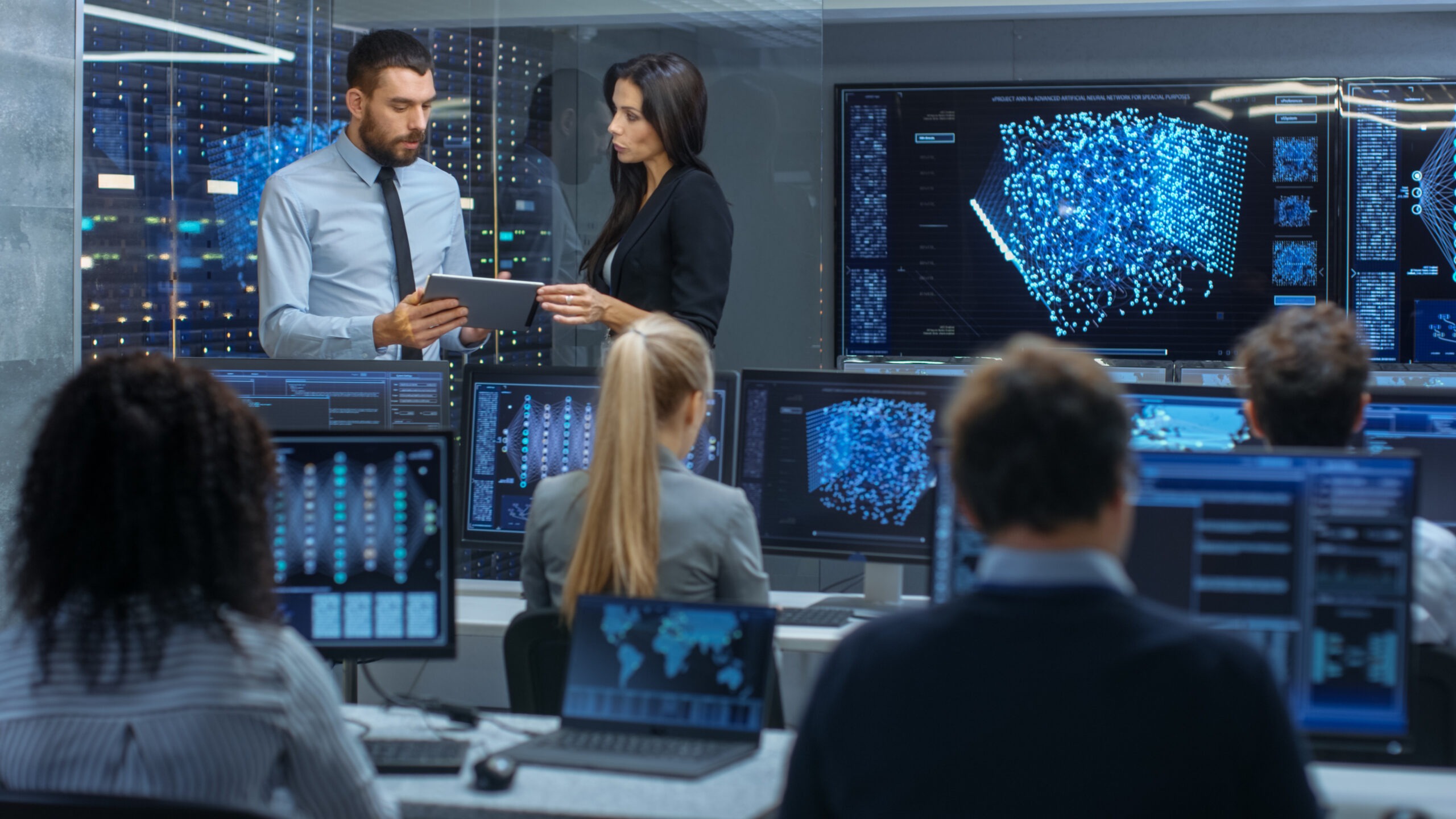 Professionals collaborate in a high-tech data center, analyzing complex data visualizations on multiple screens while discussing insights on a tablet.
